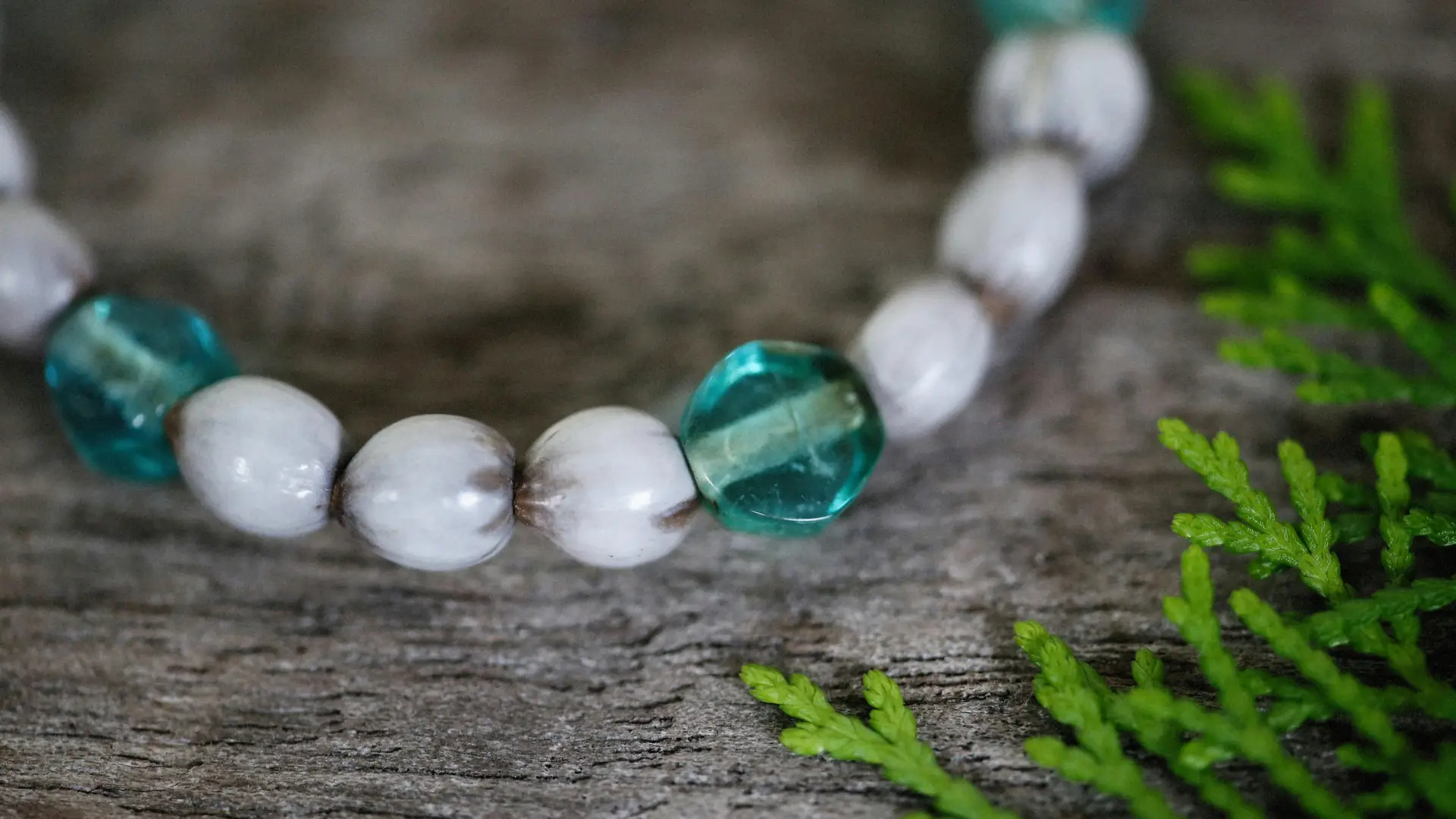 A close-up of a beaded bracelet with white and teal oval beads on a wooden surface, next to green fern-like leaves.