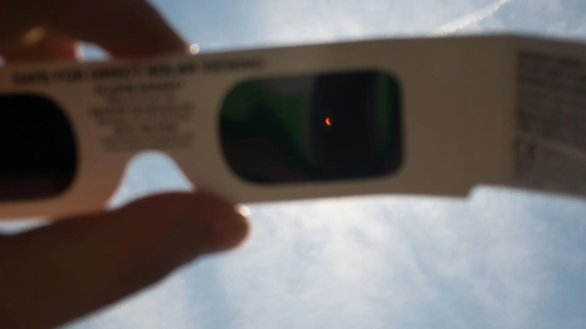 A partial solar eclipse is visible through eclipse glasses held up to a sunny sky.