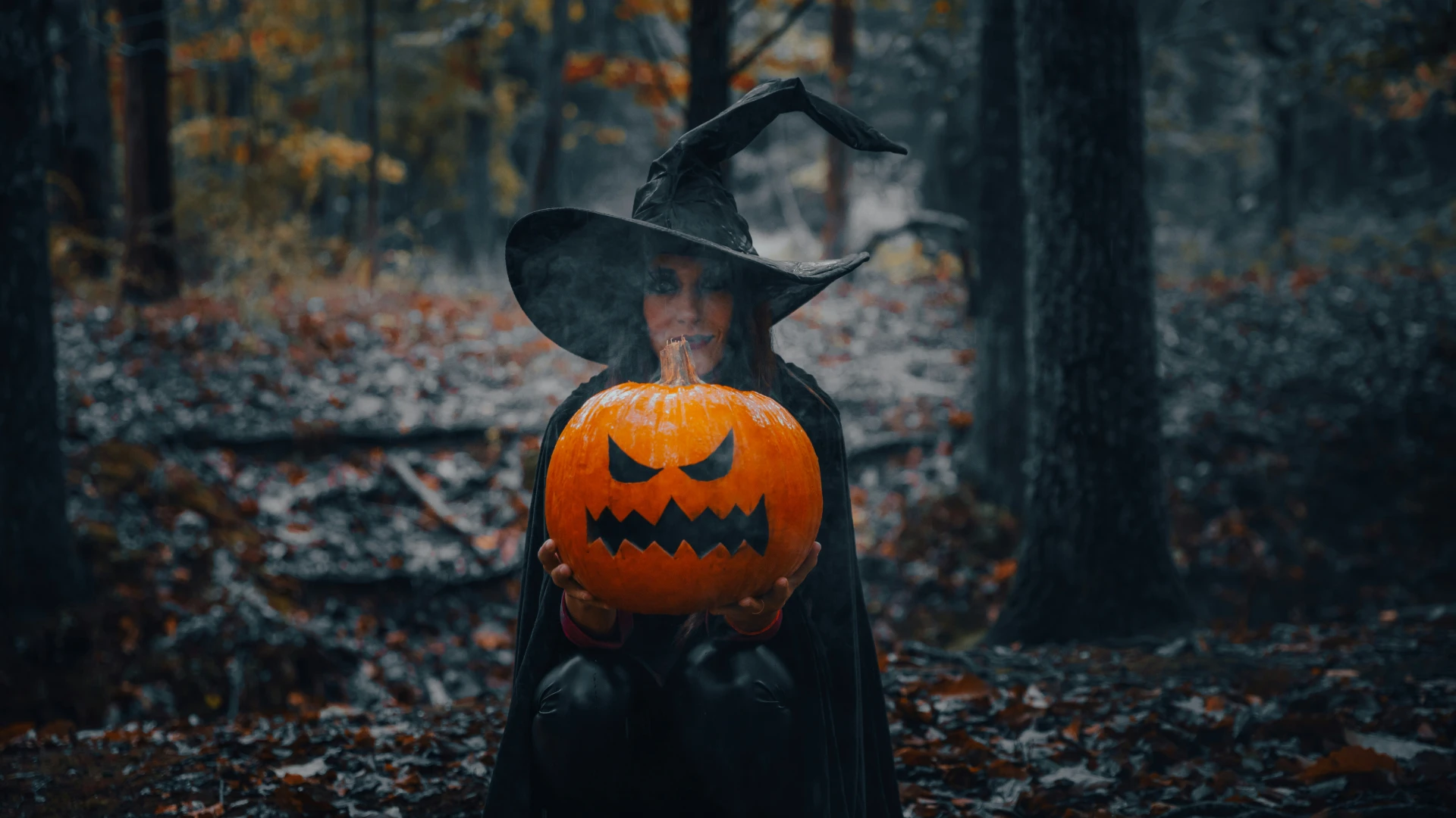 Woman stands in woods in black witch robes and hat holding large jack-o-lantern