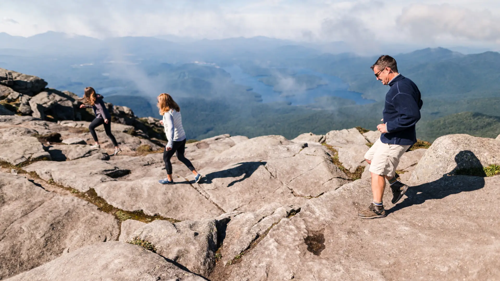 A man and two women in summer clothing walk across a rocky mountain summit on a sunny day.