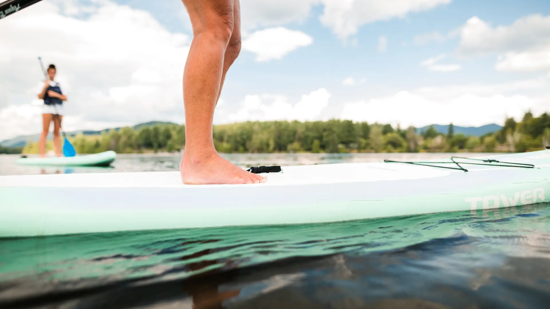 Person's feet and legs seen standing on a white paddle board with a second person paddling a paddle board in the background