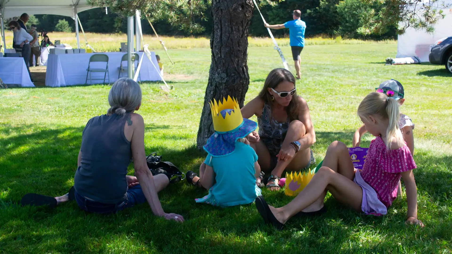 A woman sits under a tree eating snacks with her young kids.