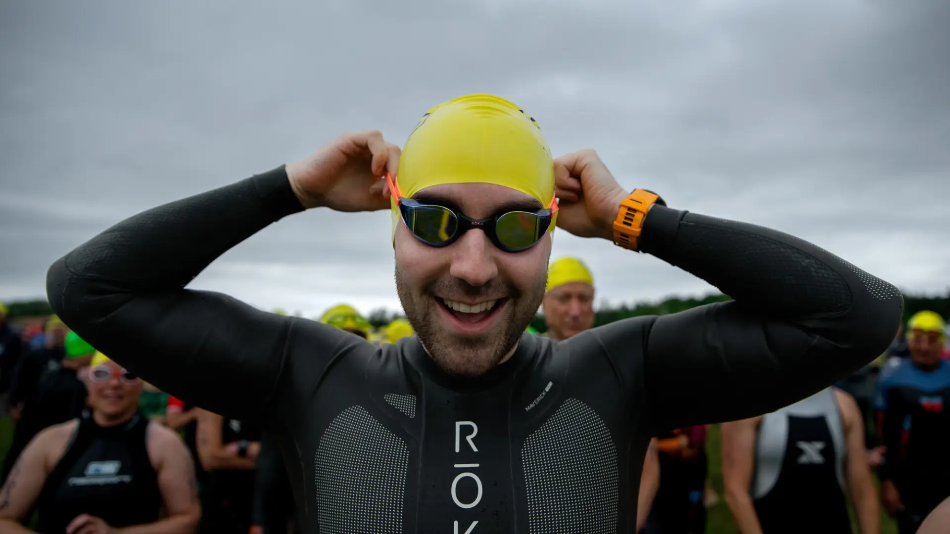 A smiling triathlete in a black wetsuit adjusts his goggles and yellow swim cap while preparing for the start of the race.