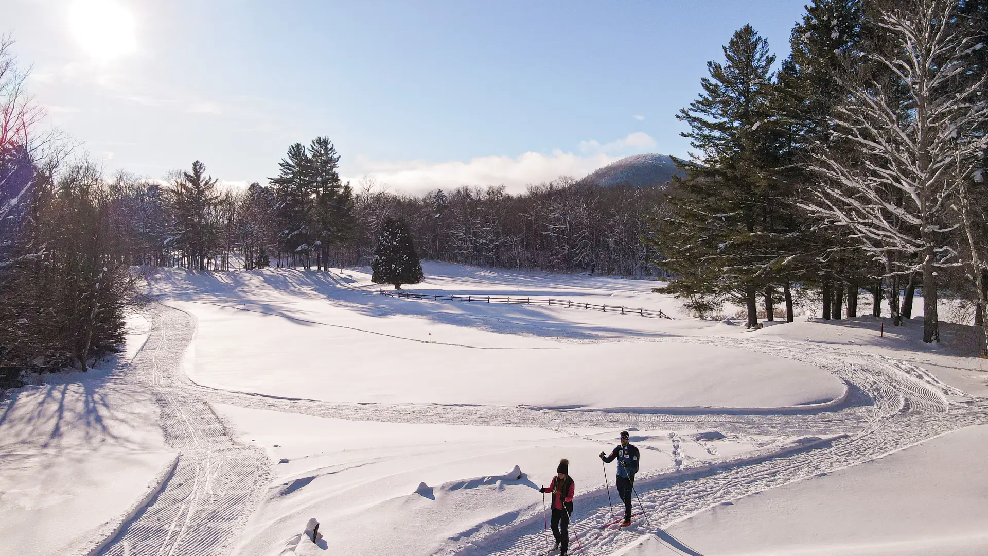 A man and woman cross-country ski on an open meadow, surrounded by trees, with a mountain in the background.