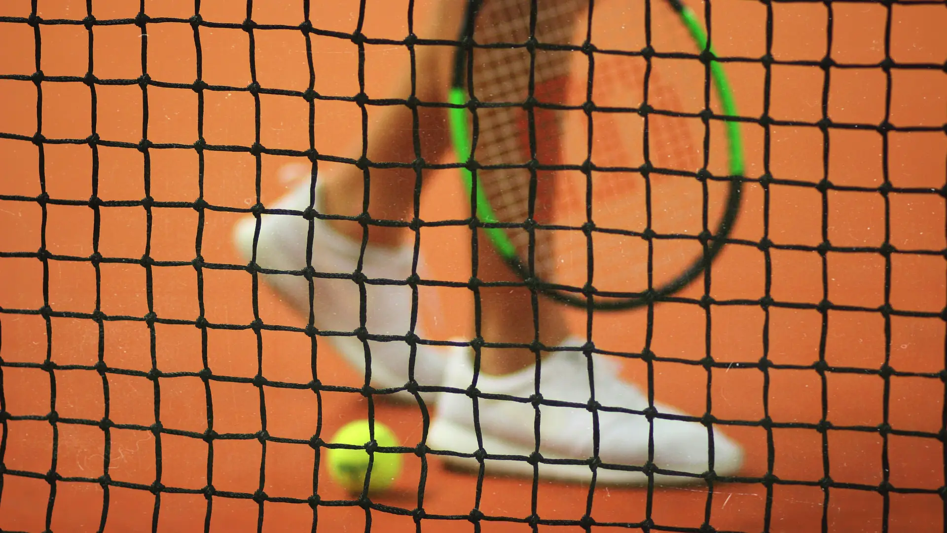 Person's feet in white tennis shoes, racket, and tennis ball seen on clay court through tennis net