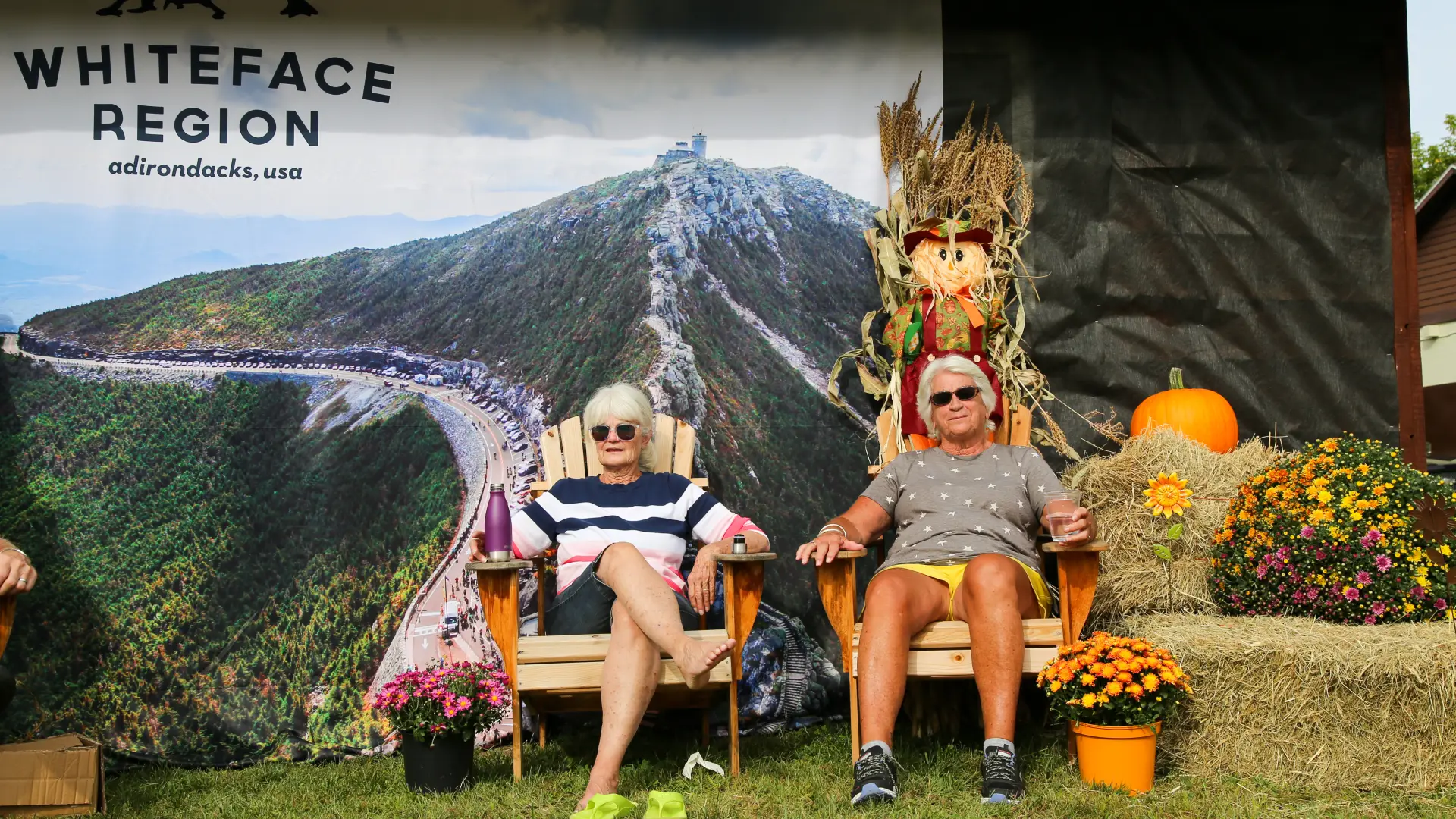 Two women sit in wooden Adirondack chairs surrounded by pumpkins, mums, and hay bales in front of a large mural image of Whiteface Mountain