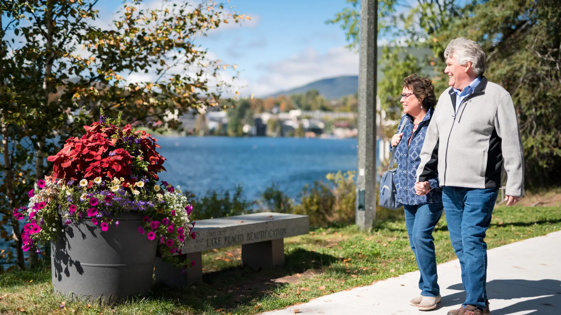 Two people walk on a sidewalk in Lake Placid village, with views of Mirror Lake in the background