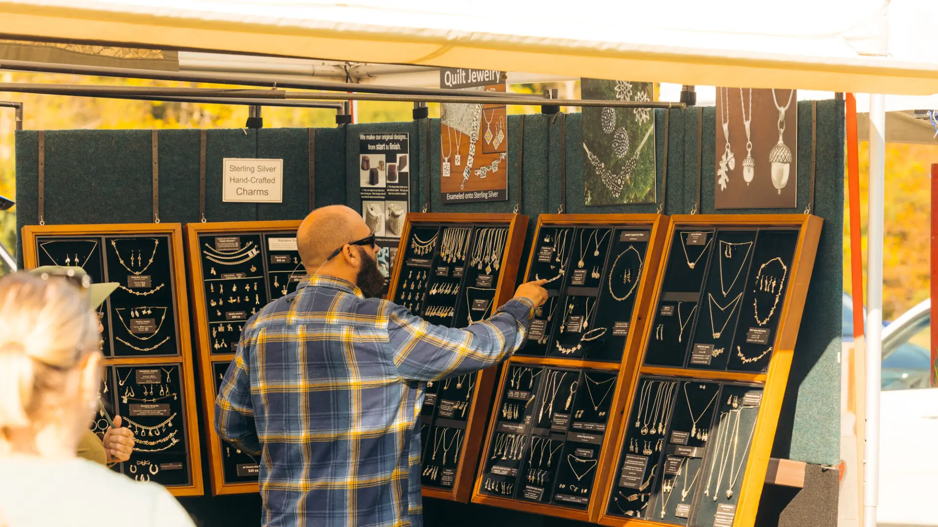Man wearing flannel and hat browses displays of silver jewelry in farmers market booth