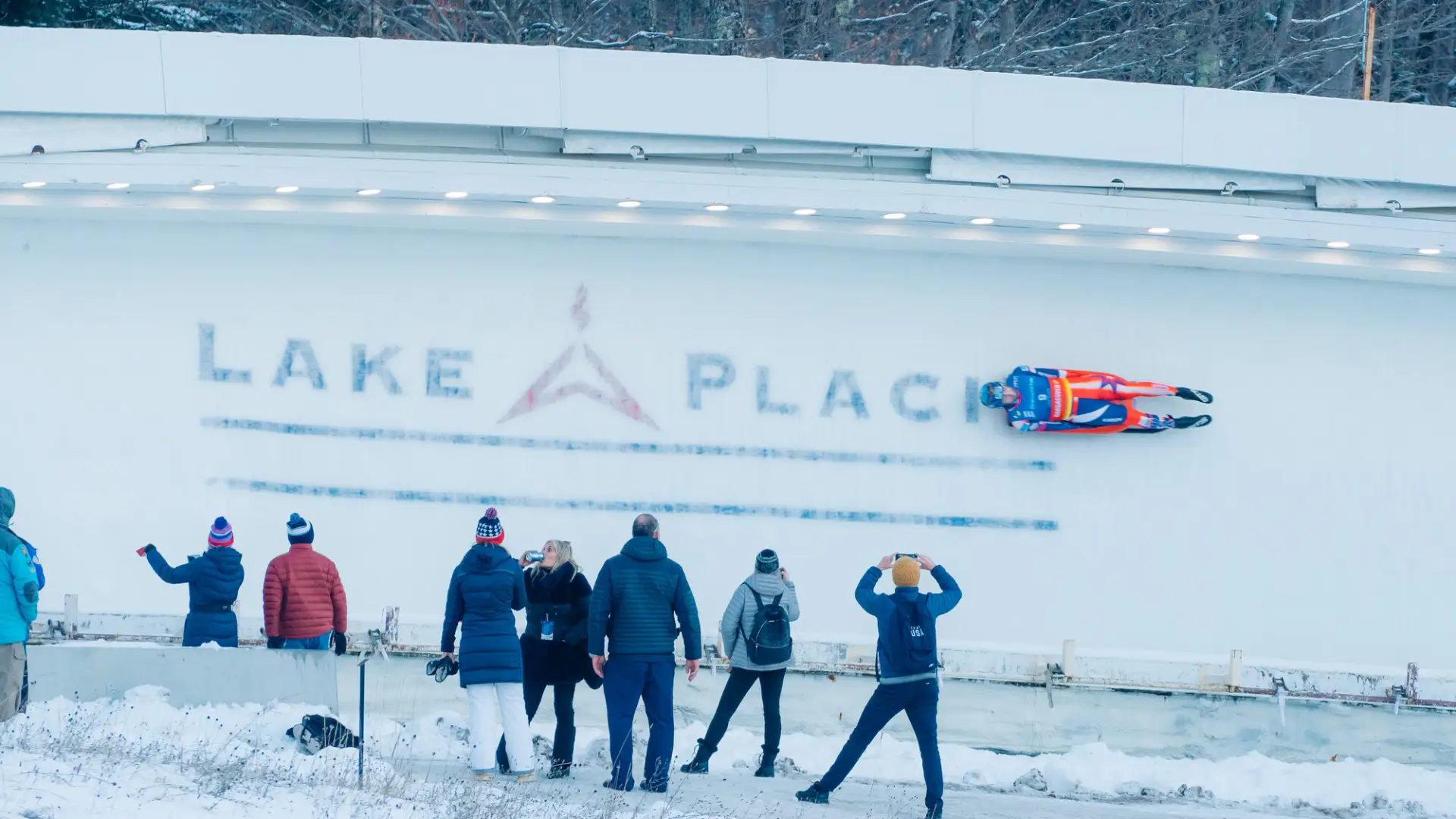 Luge racer rides down track with Lake Placid in ice while crowd looks on