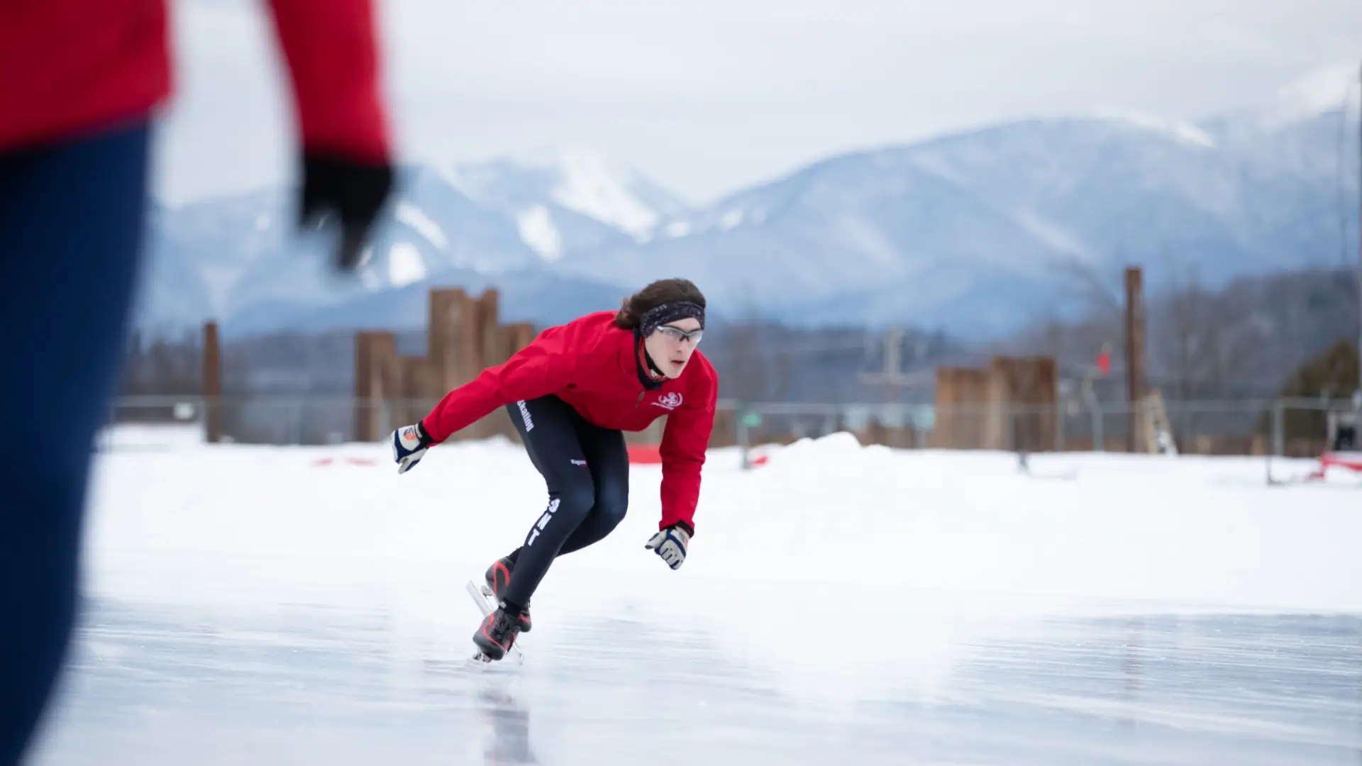 Skater in red coat crouches and skates on outdoor ice skating oval with mountains in the background and snow on the ground