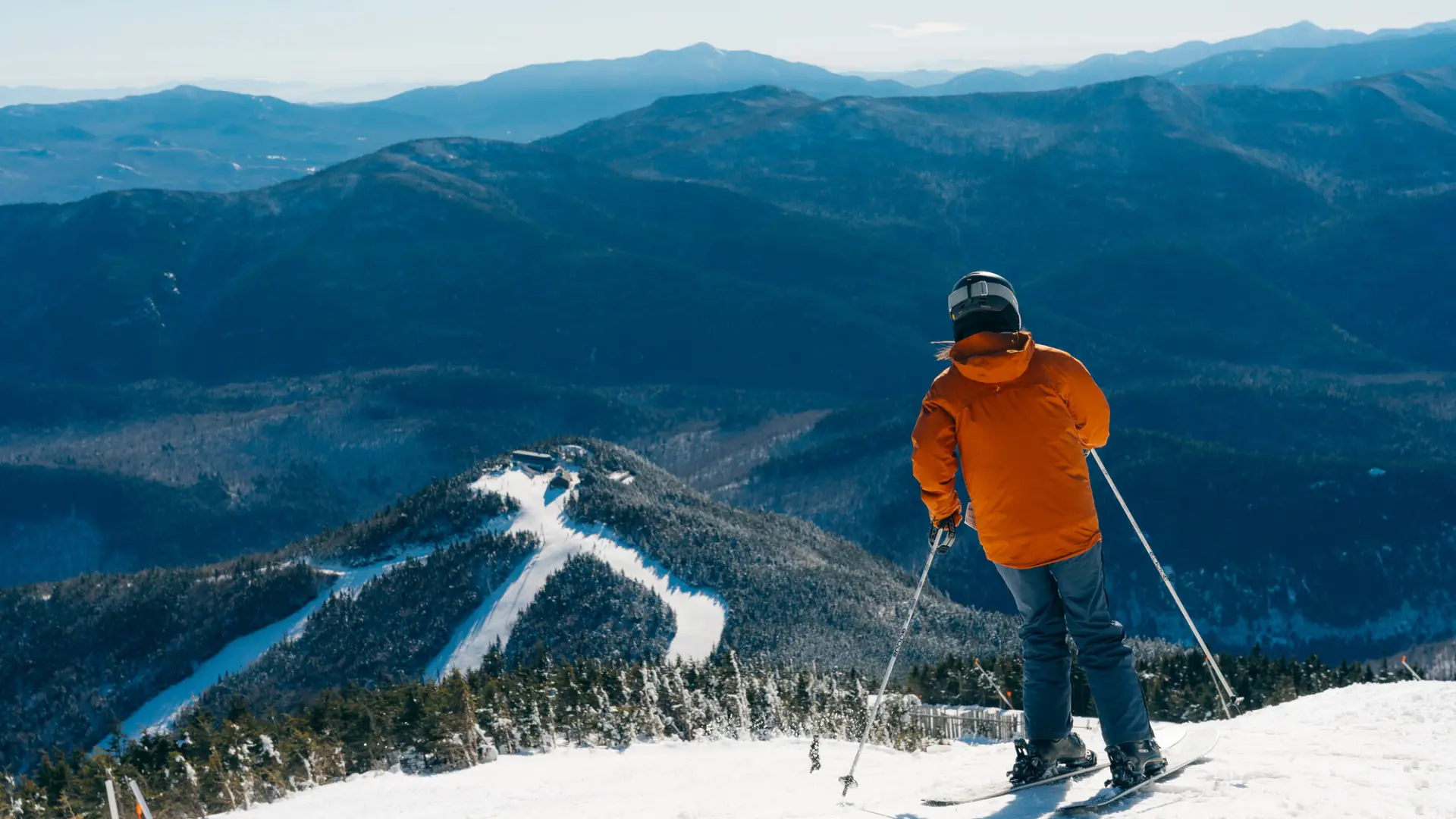 A skier in an orange jacket stands at the top of a snow-covered mountain, looking out over a range of blue-tinted peaks.