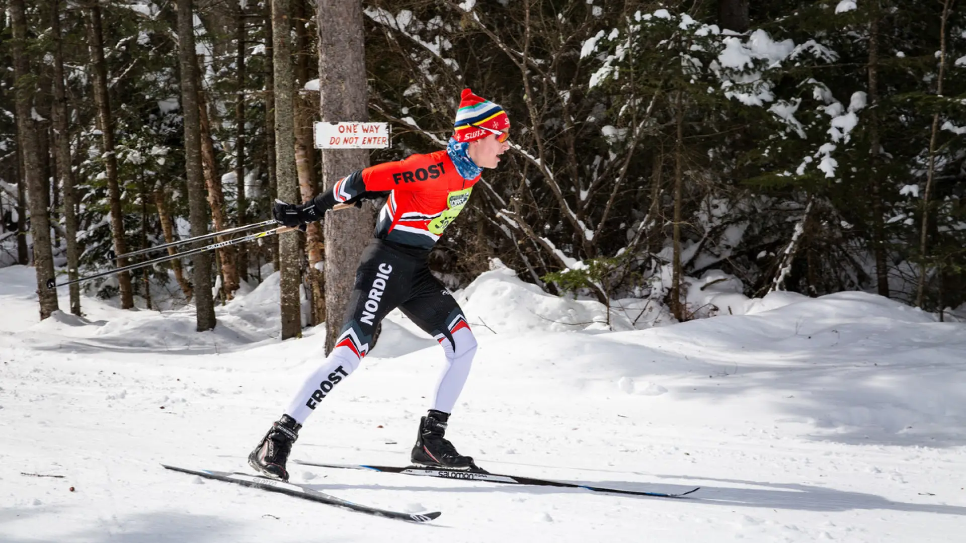 A single nordic skier passes by the camera from left to right, skiing on a snow-covered trail through a pine forest. They wear red, black, and white ski race gear and a yellow race bib on their chest
