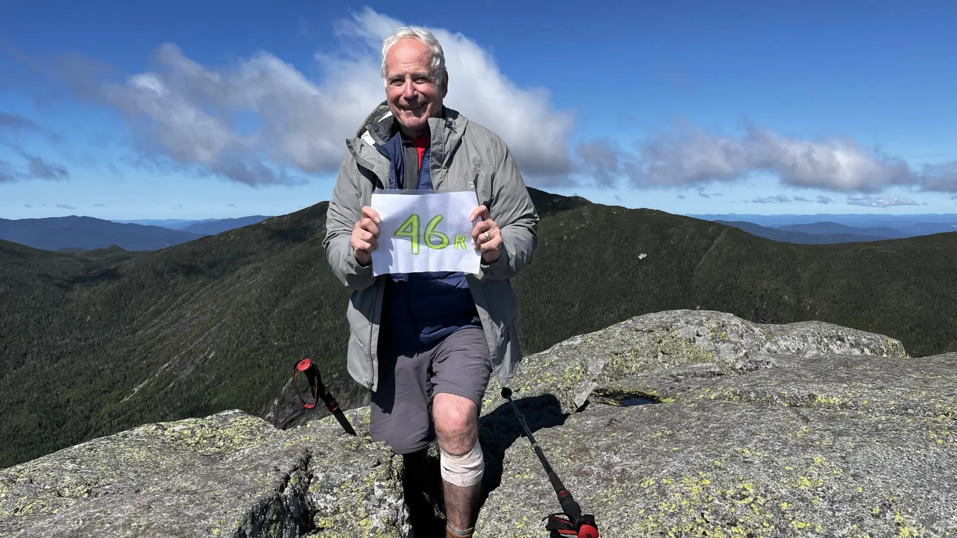 Older Gentleman stands atop a mountain peak holding a sign that reads '46er!'.