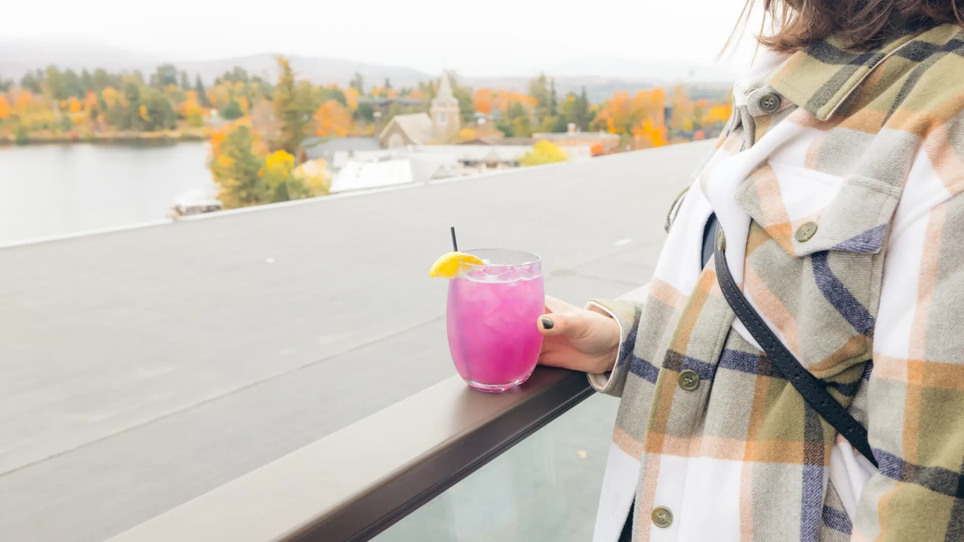 Woman in light flannel coat and painted fingernails holds glass of purple cocktail on rooftop bar overlooking Lake Placid in fall