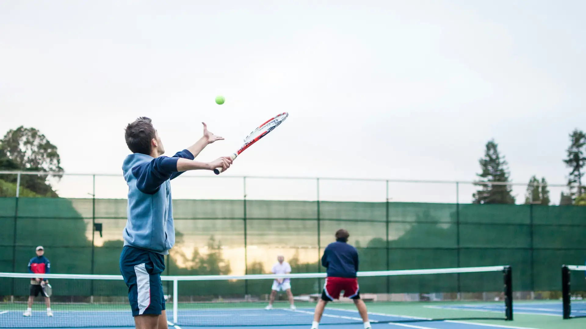 Man serves a tennis ball to an opponent on a tennis court beside another court where two players volley