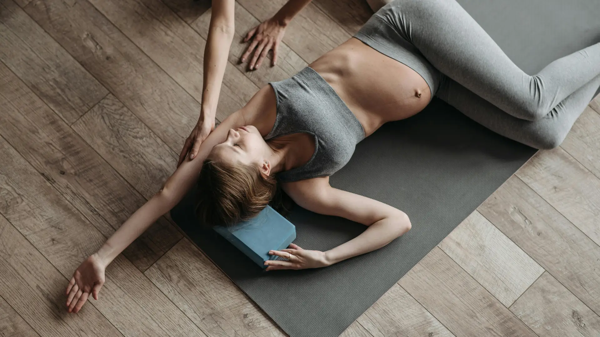 Pregnant woman in athletic clothing stretches on grey yoga mat on wooden floor with another person's arms helping deepen the stretch