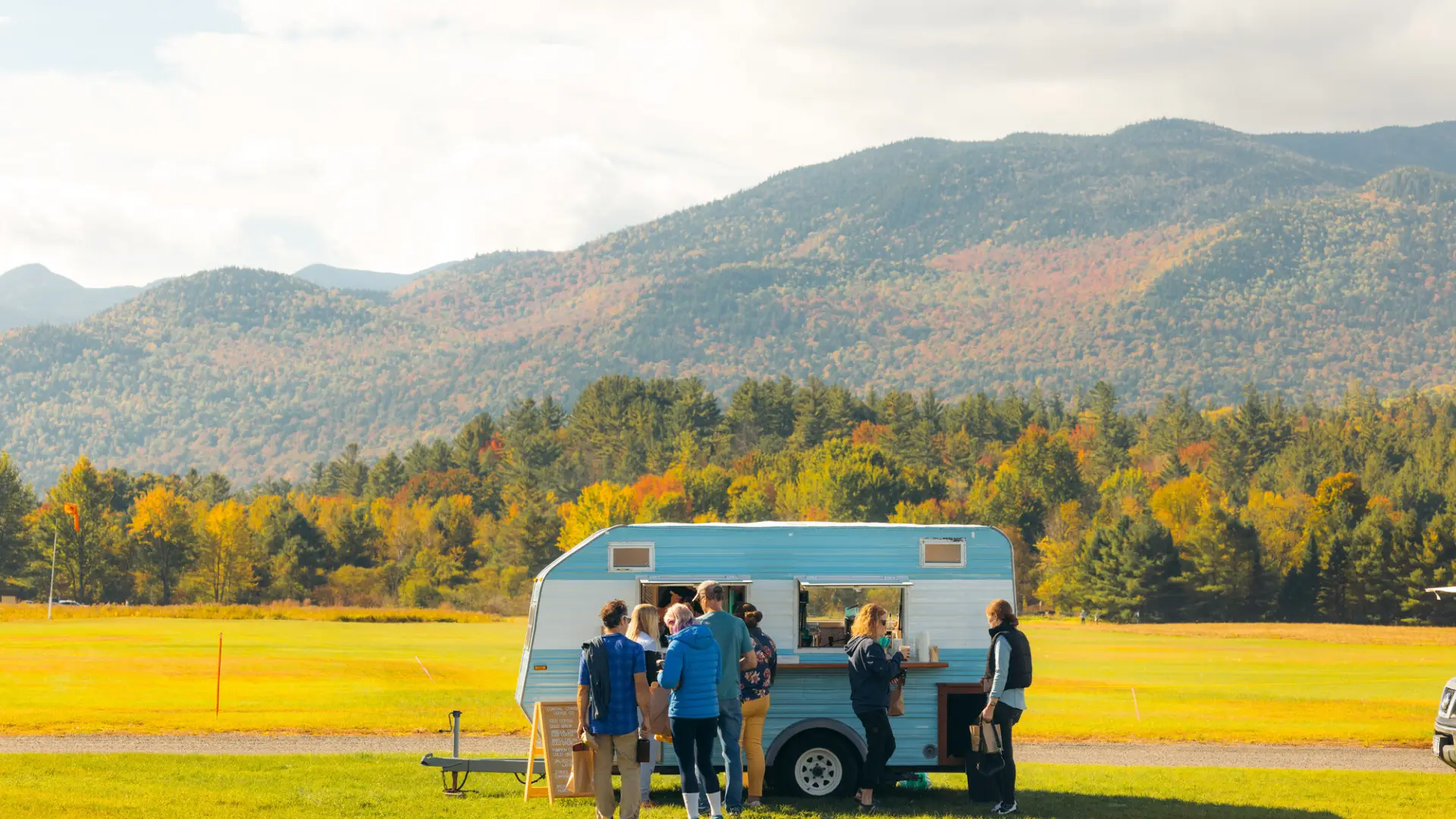Blue and white camper trailer parked in field with mountains in background and people ordering coffee at window