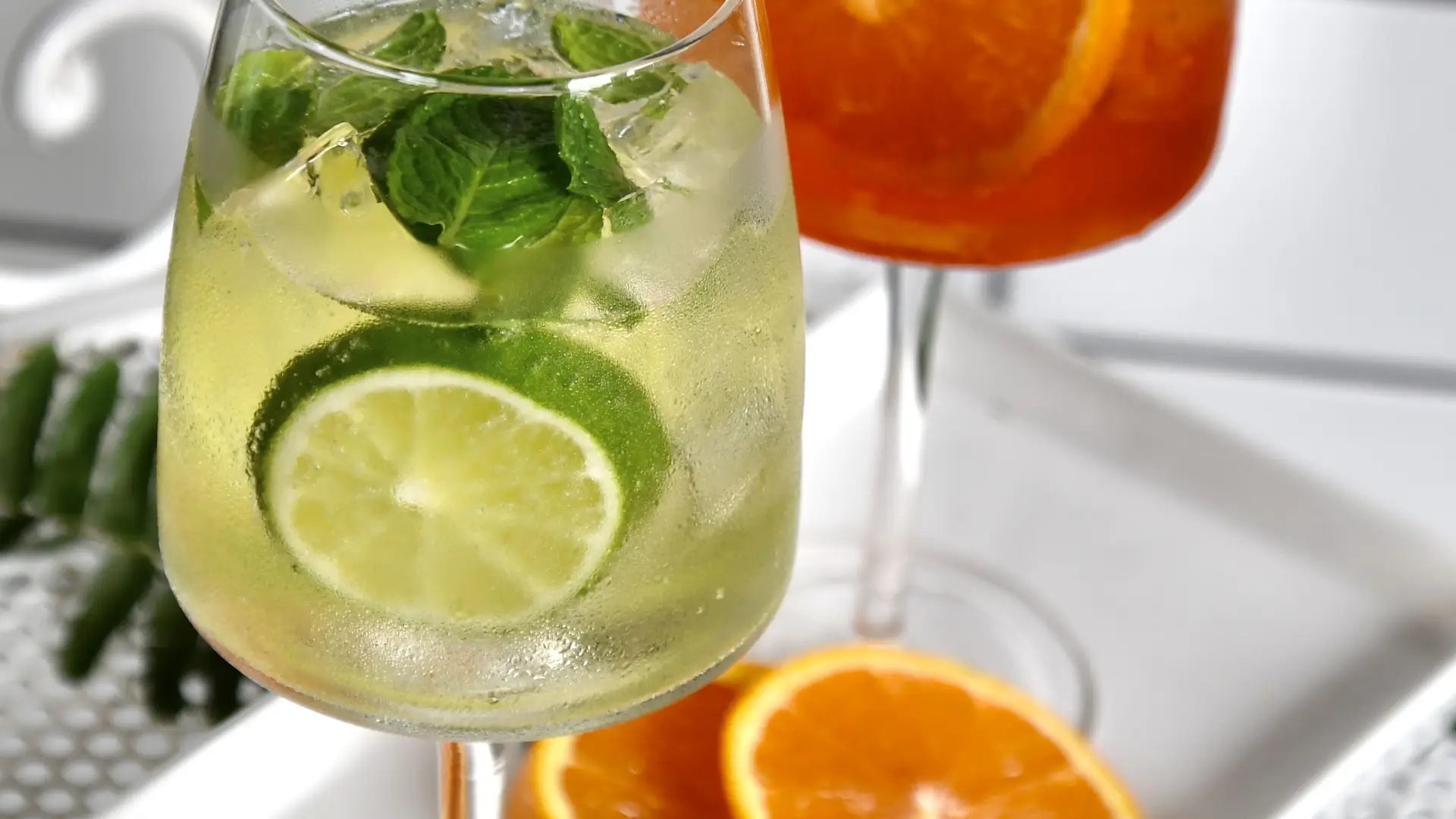 Two cocktails that look like a mojito and an Aperol spritz with a rosemary sprig on a white plate with citrus garnishes