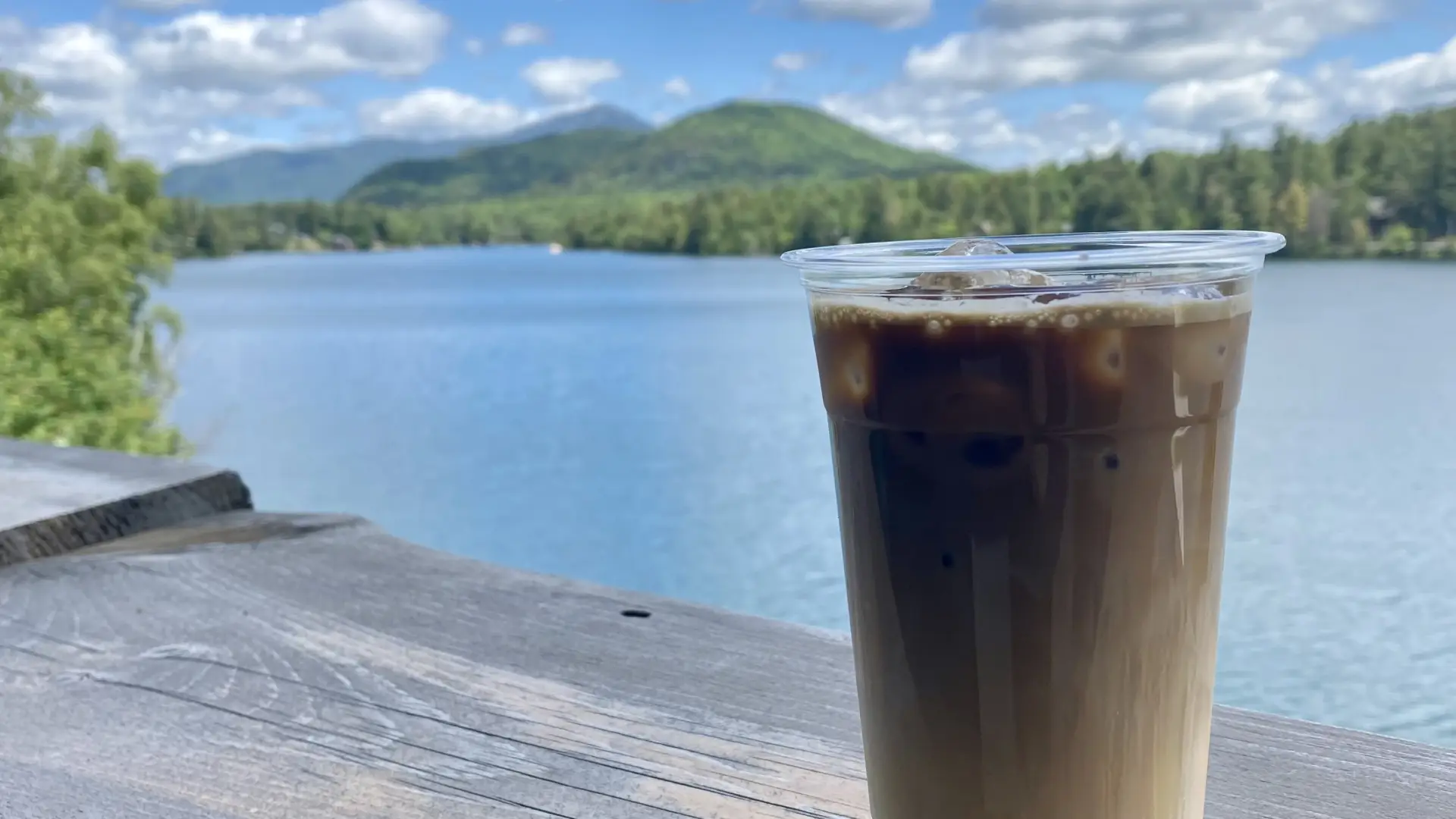 A cold coffee sits on a railing overlooking a sunny lake and mountains.