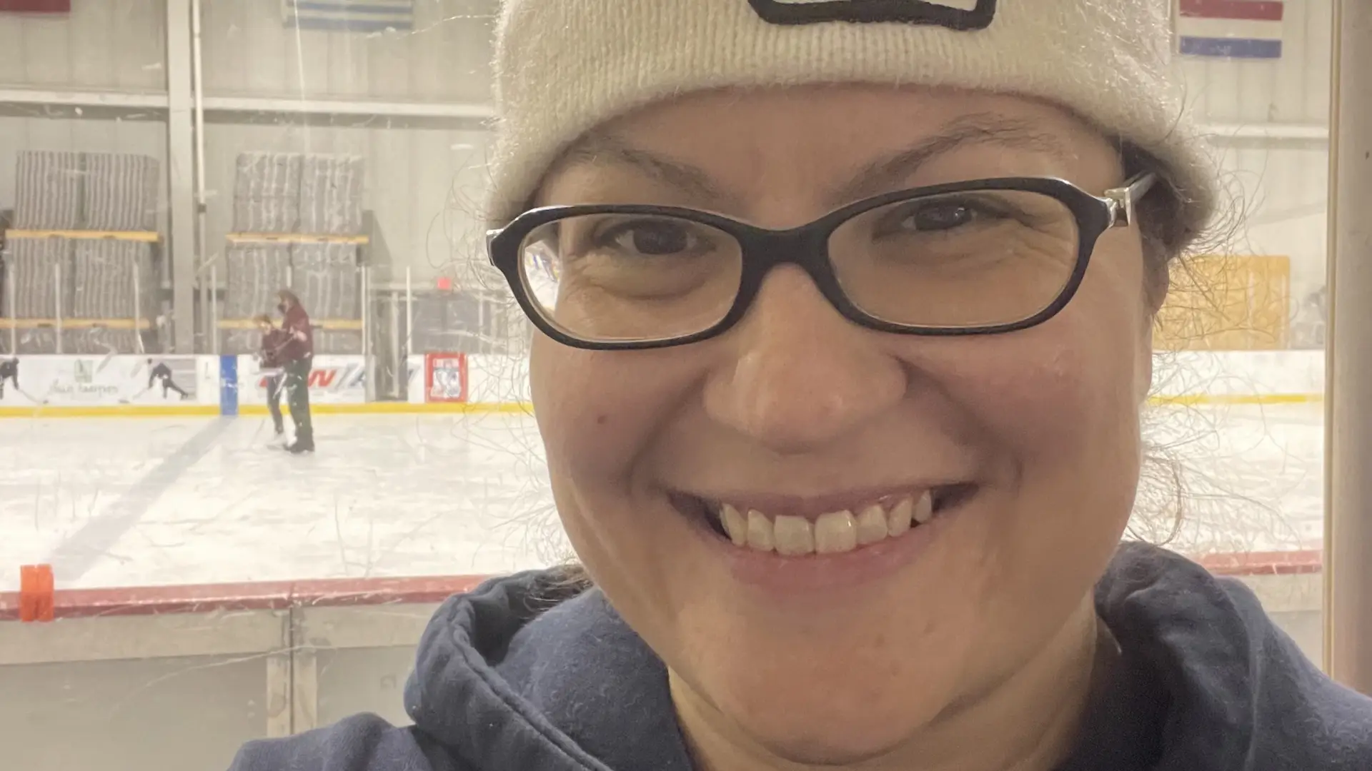A woman in a 1980 Lake Placid Olympic winter hat smiles in front of an indoor skating rink.