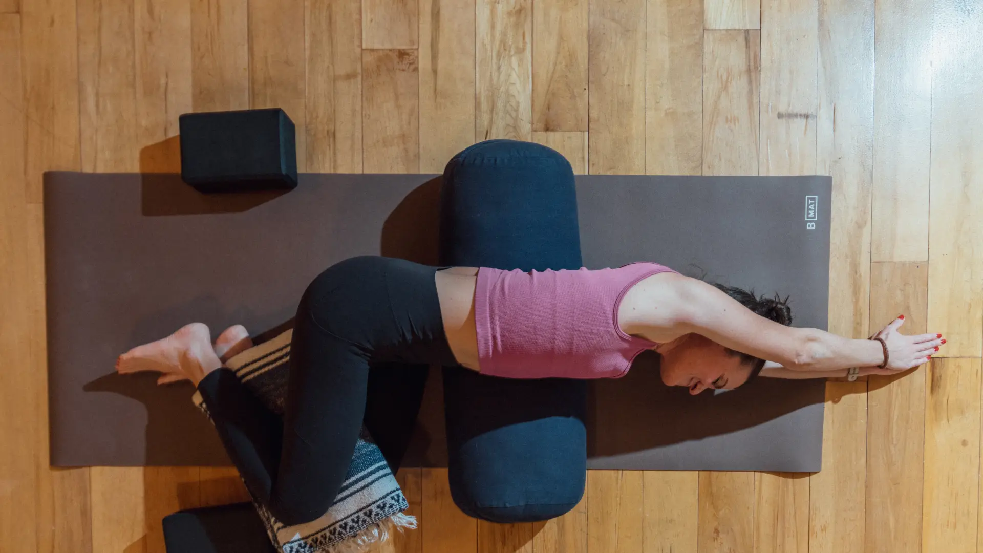 Woman in pink tank top and black yoga pants lays on side on black yoga mat on wooden floor with legs bent, a blanket between her knees, and her arms extended above her head
