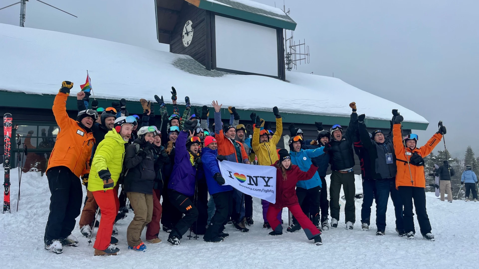 A large group of skiers and snowboarders in winter gear cheer and wave while holding an "I Love NY" LGBTQ+ pride flag.