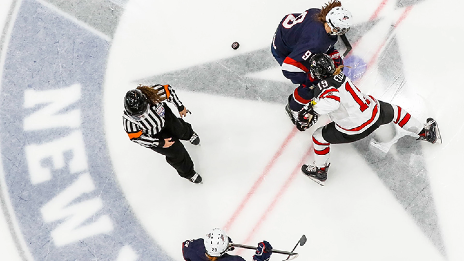 Two female hockey players face off on ice with another playing and a referee looking on