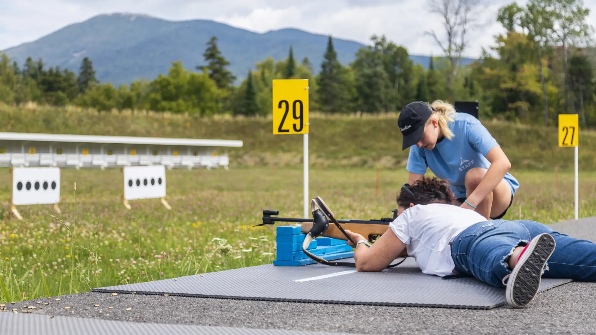 ORDA staff instructor kneels next to woman lying on biathlong shooting platform with biathlon rifle at range in summer