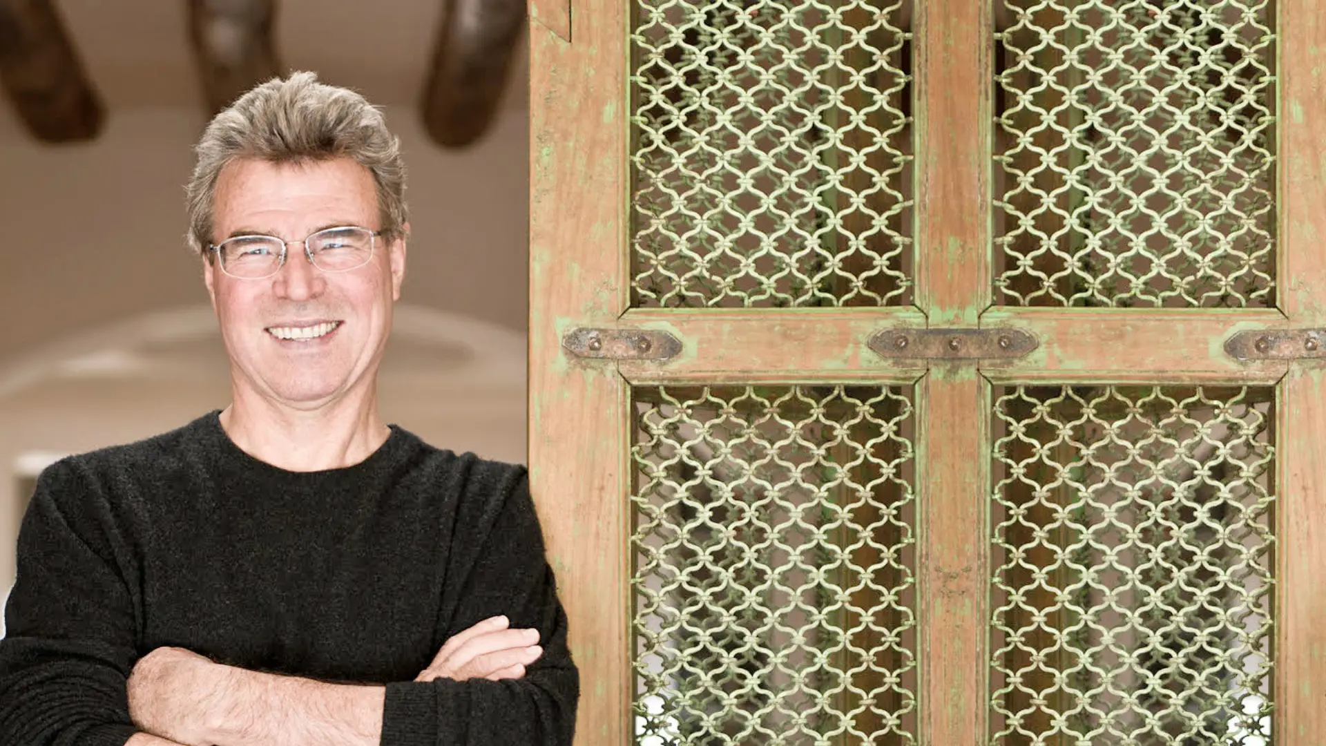 Steve Thomas smiles next to a decorative, green-painted wooden screen with a patterned metal grille.