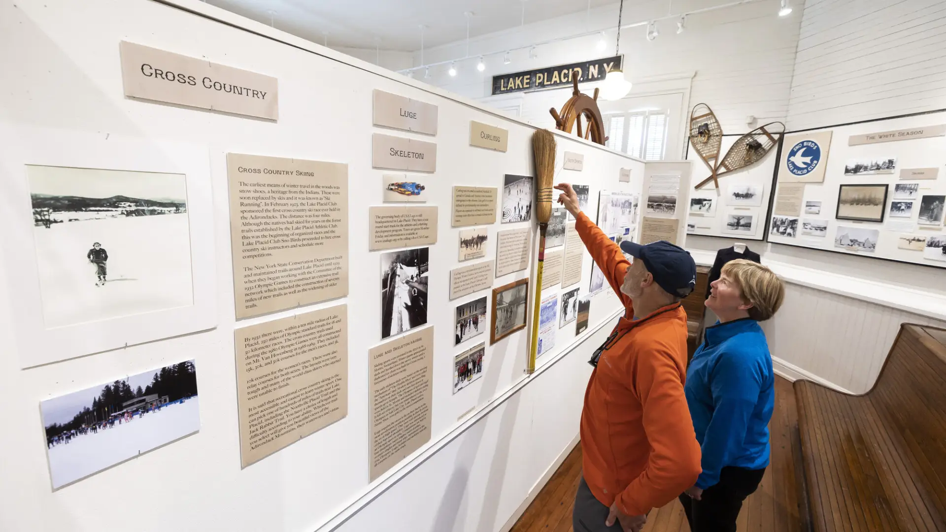 Senior man wearing orange sweater and hat stands next to senior blonde woman with blue jacket, the man pointing at a straw broom that is part of a historical display wall