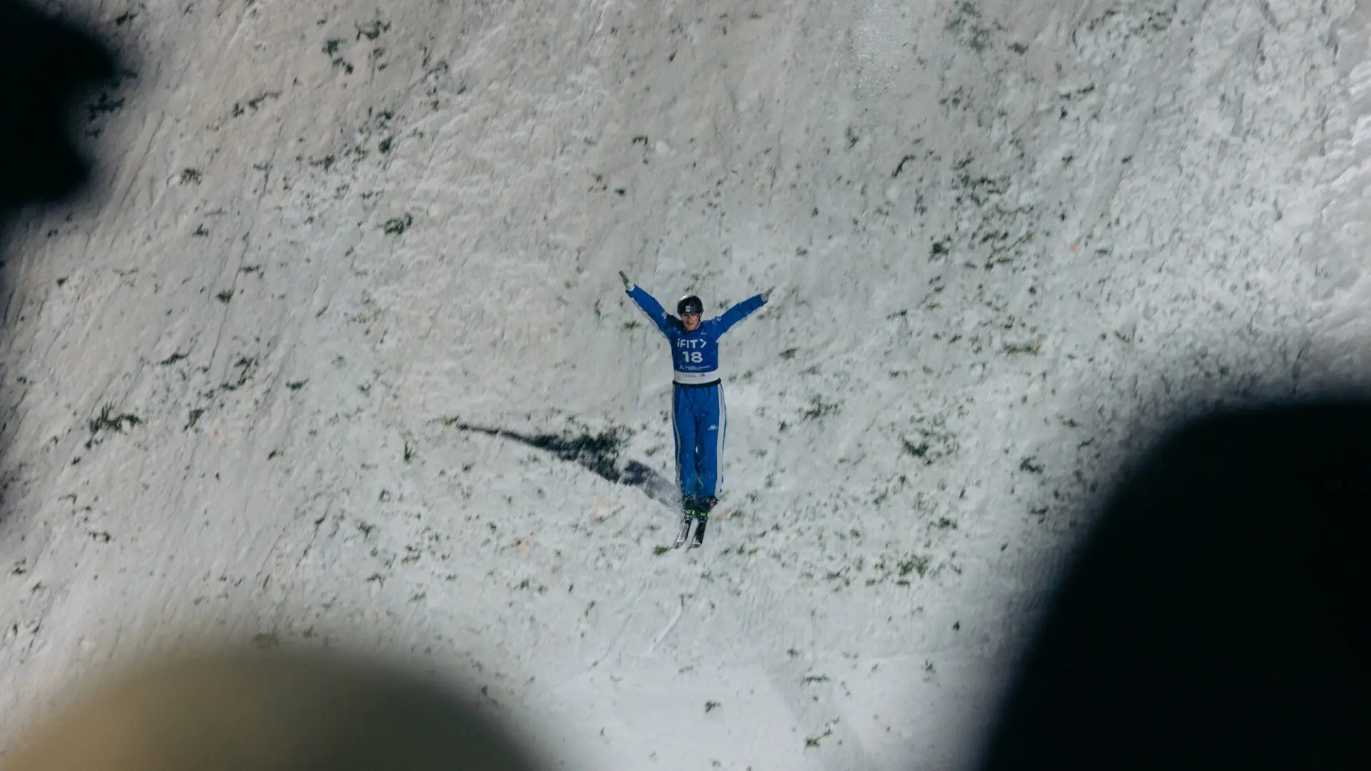 Freestyle aerial skier in a blue snowsuit lands on snowy hill sprinkled with pine bows. Their arms are extended out and up, while their legs  and skied feet are tight together