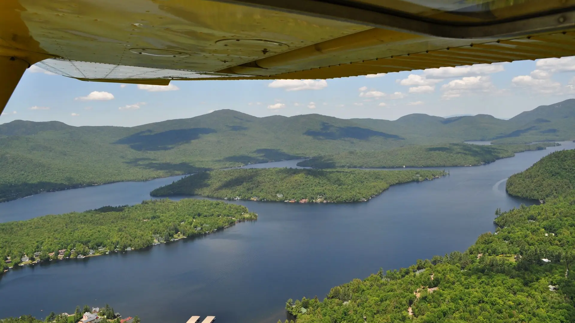 Looking down on green forests, a forested island, and blue lake from under a small yellow airplane wing.