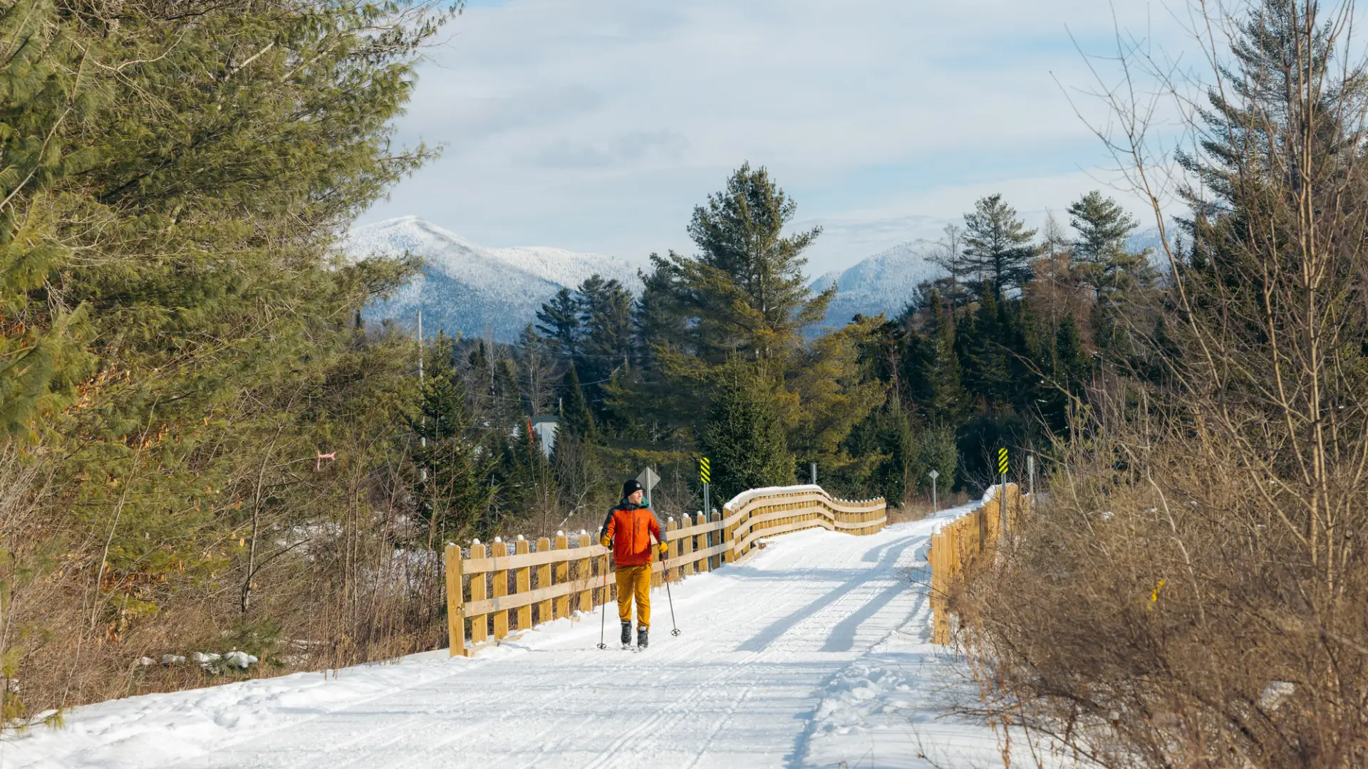 Person in red snow coat cross-country skis on Adirondack Rail Trail near bridge with snowy mountain seen in background