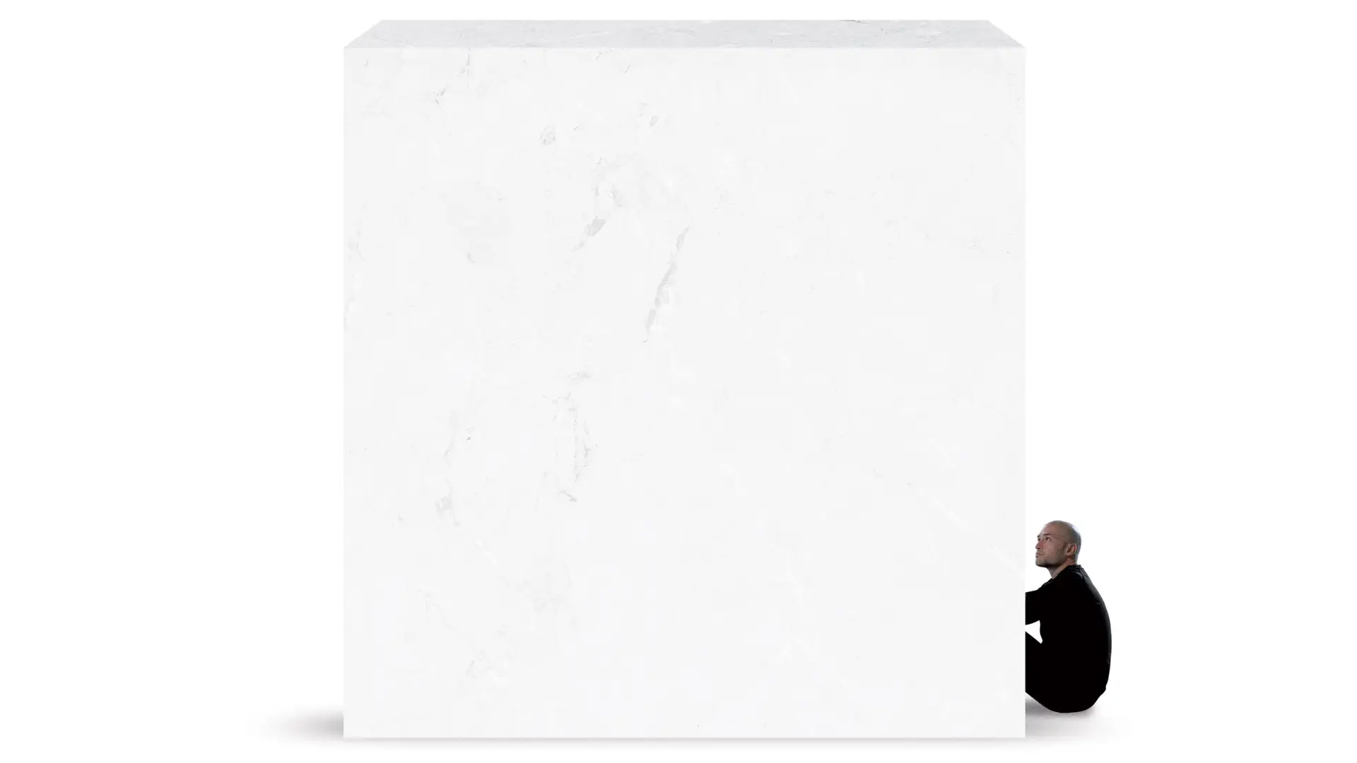 A small person in a black outfit sits tucked against the base of a massive, white marble cube that towers over them.