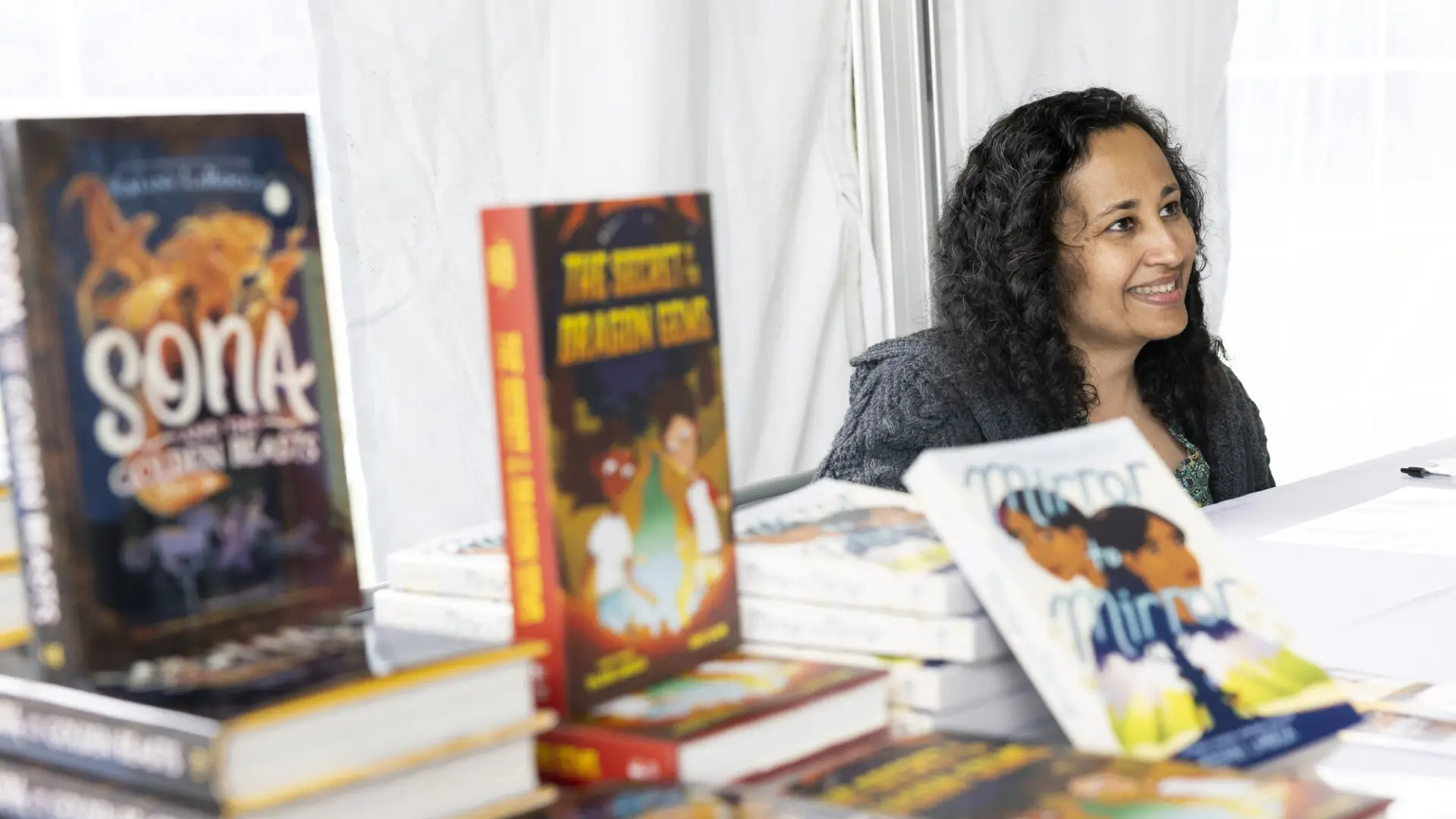 Female author seen sitting and smiling at someone off camera from behind a stack and display of children's books