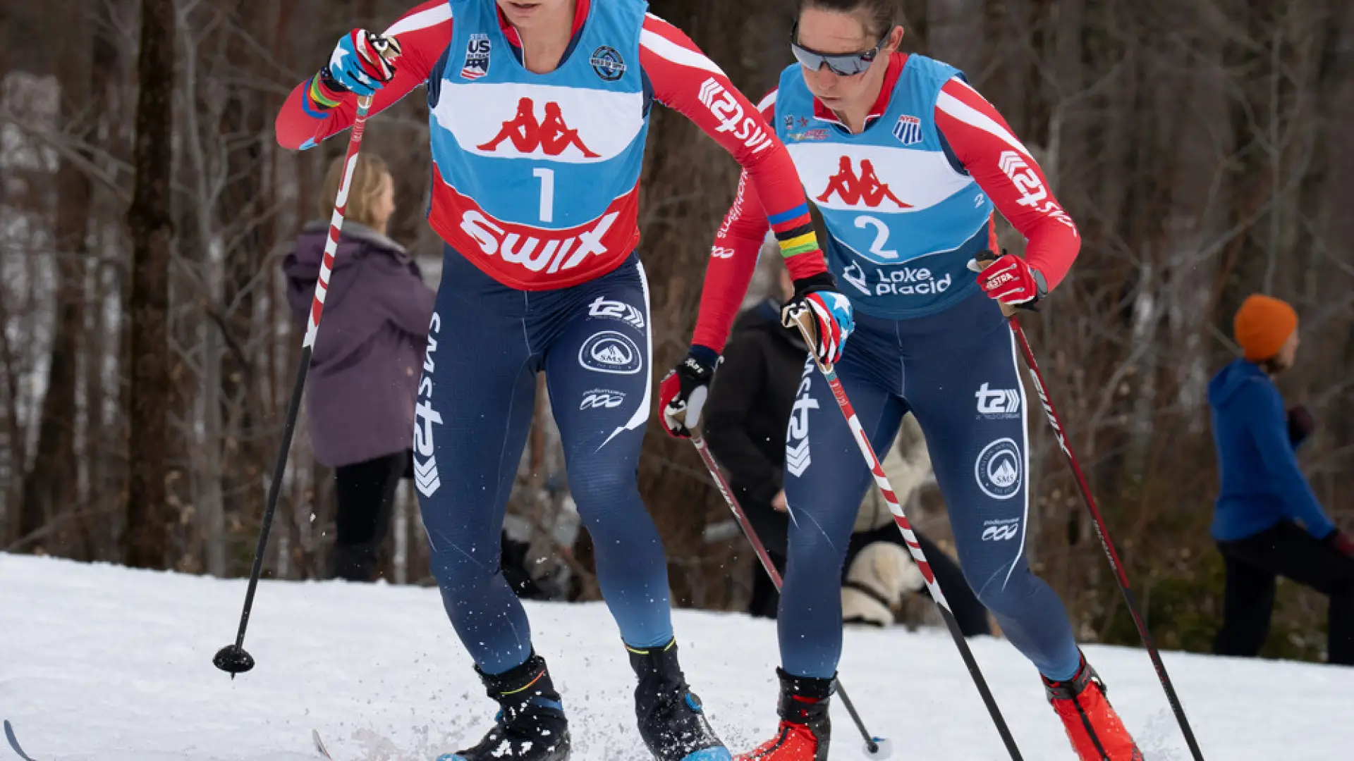 The image shows two female cross-country skiers racing on snow. 