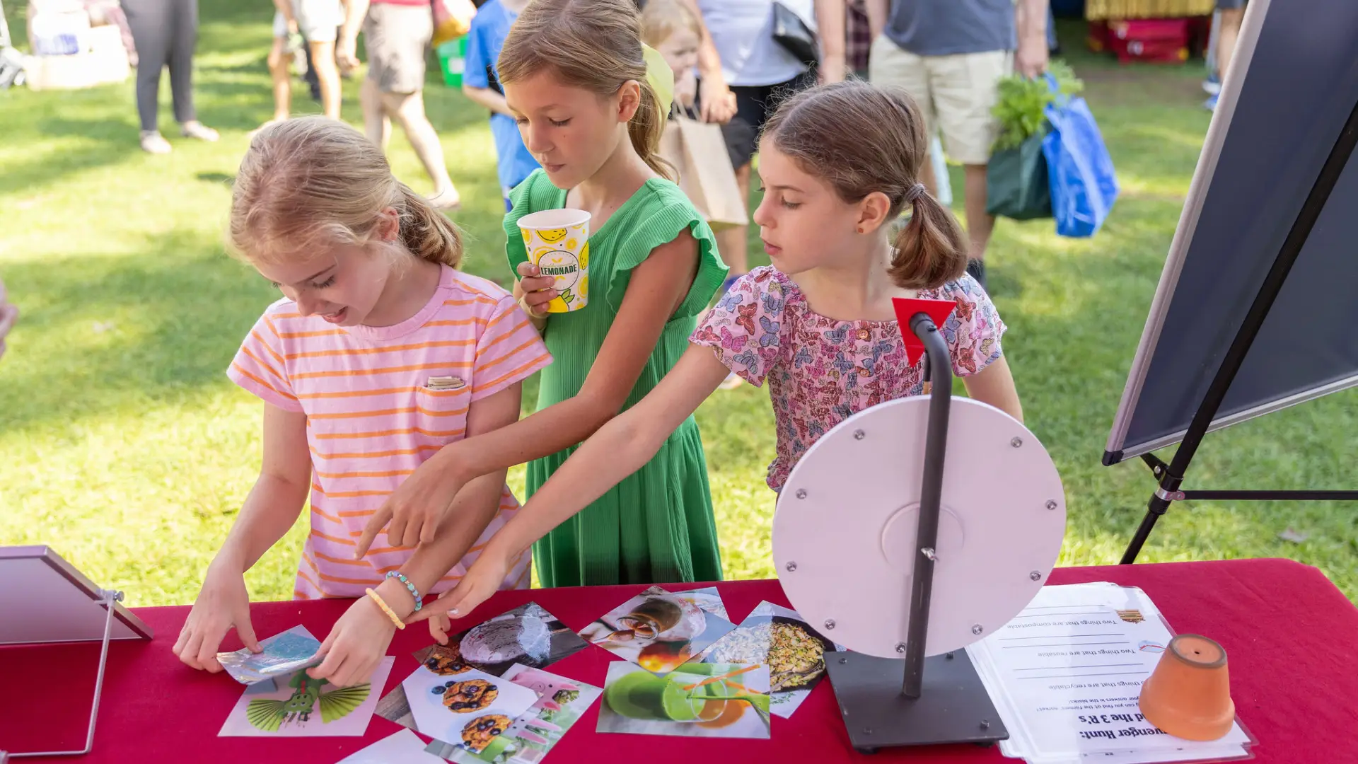 Three girls at a table with a prize wheel, looking at pictures of food and other items.