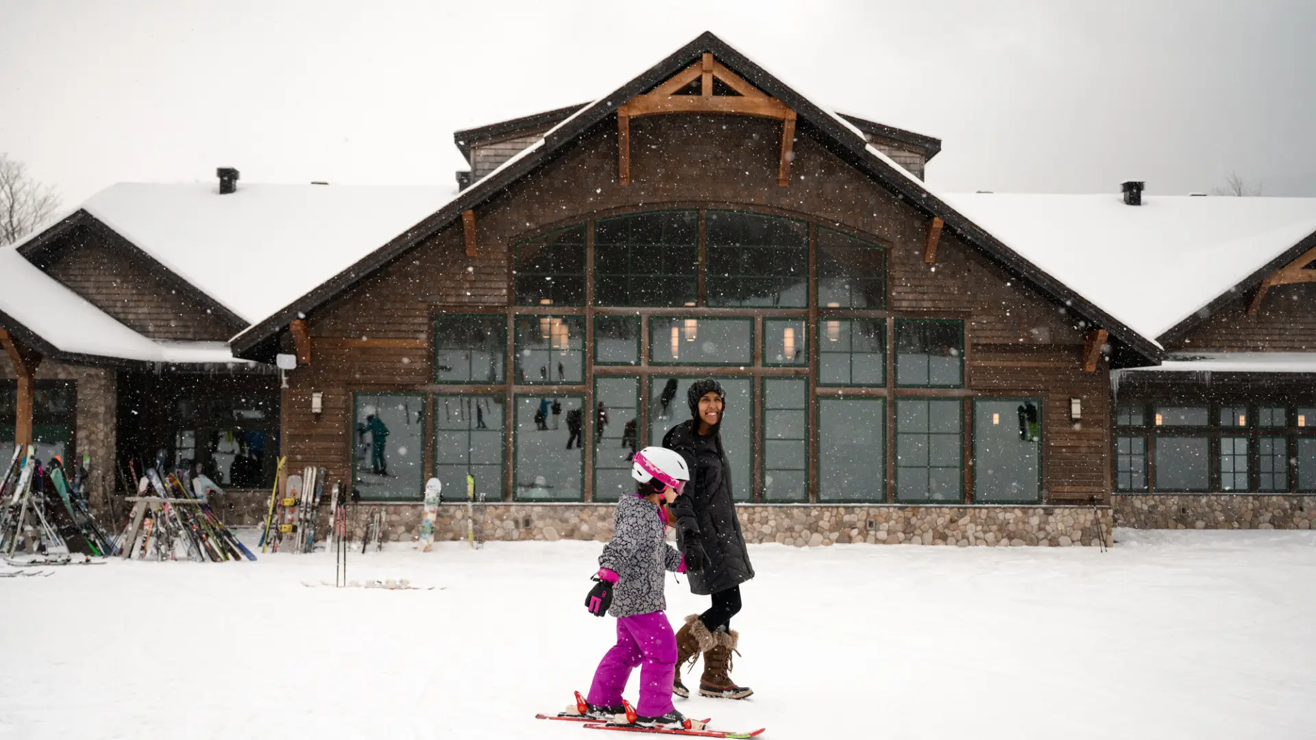 A woman in a black parka and a young girl in bright purple snow pants and a pink helmet walk together at Whiteface Mountain.