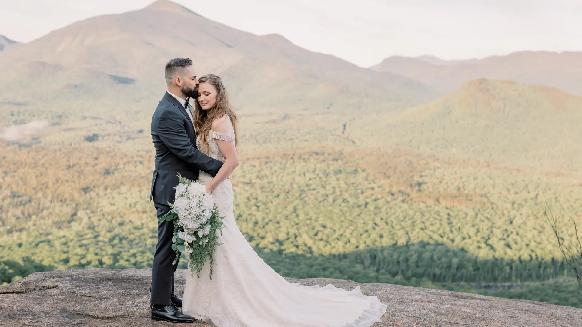 Groom kissses bride's forehead while she displays her bouquet on a rocky face with mountain views