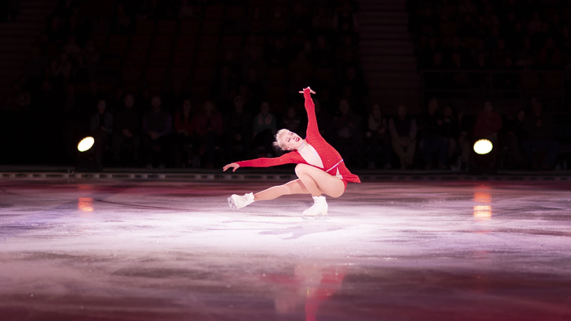Female figure skater in red dress performs a crouched move on spotlit ice in dark arena