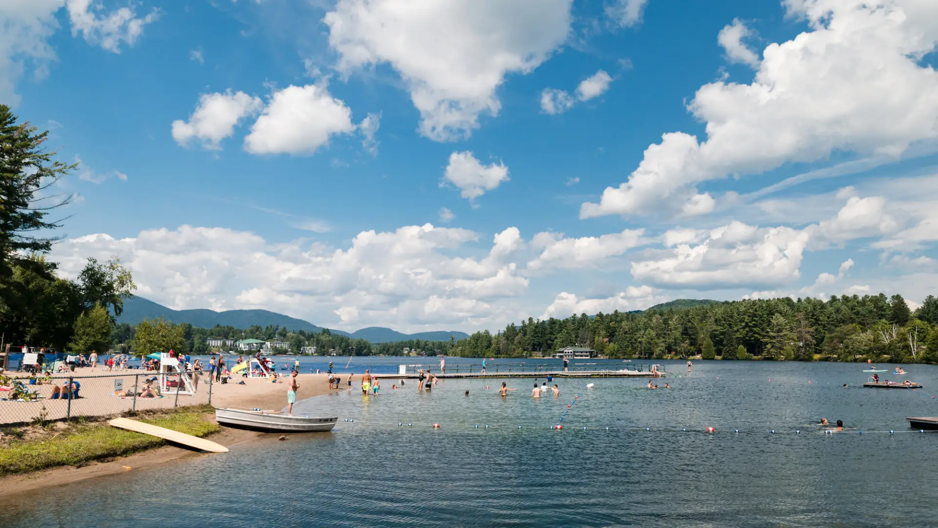 A wide view of a sparkling blue lake with a small sandy beach on the left. A dock extends into the water and swimmers are in the water. Low mountains are in the distance.