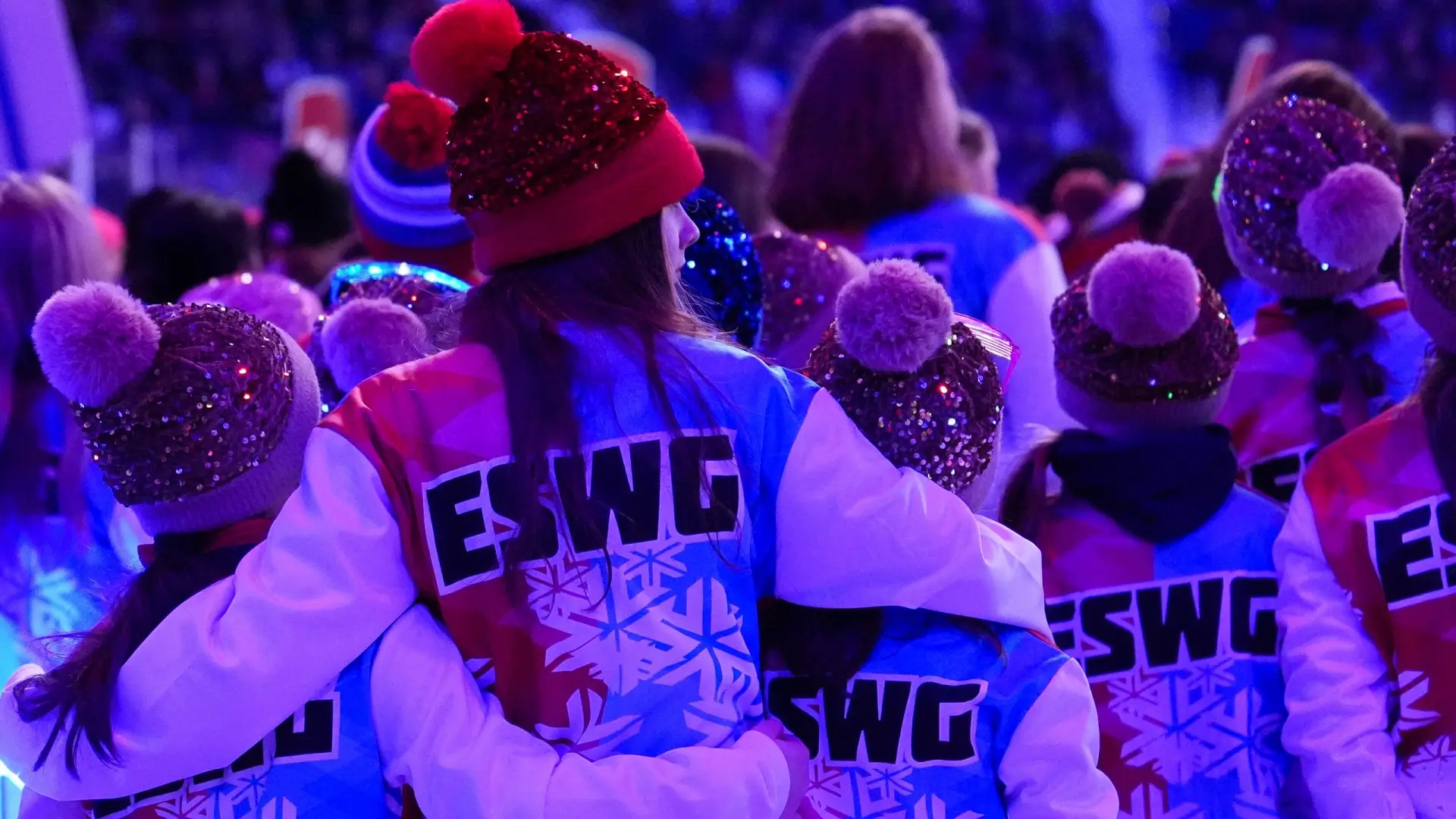 A group of people, mostly children, wearing festive red, white, and blue jackets with "ESWG" and snowflake designs, huddle together affectionately.