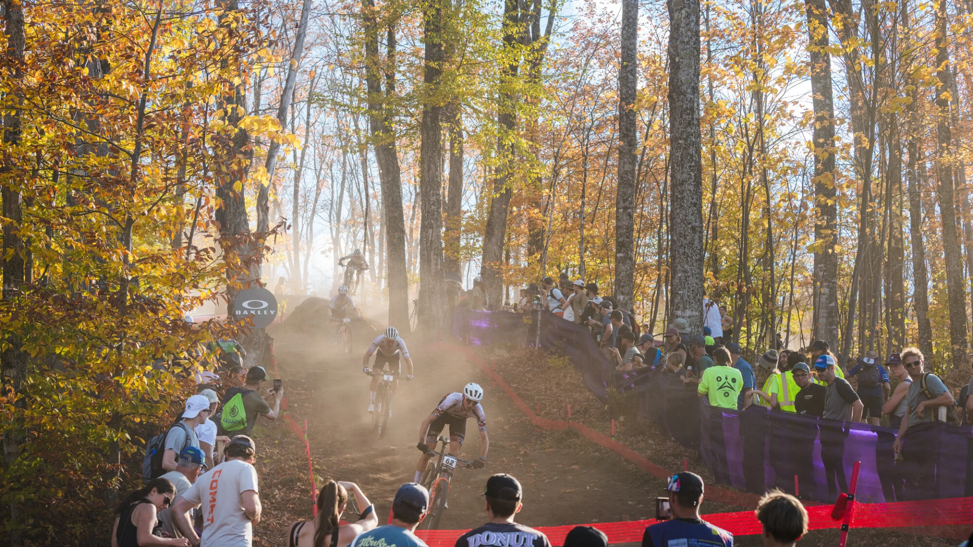 Mountain bike races ride down dirt course in forest with crowds flanking the course