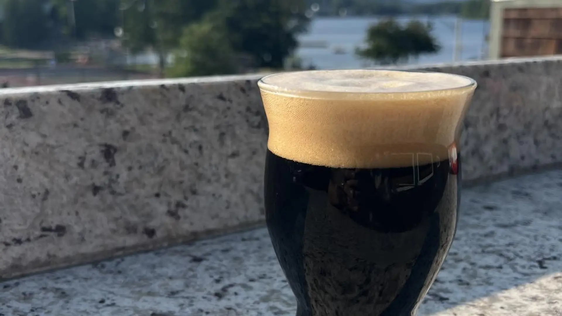 A dark glass of beer with a thick head of foam sits on a stone outdoor bar.