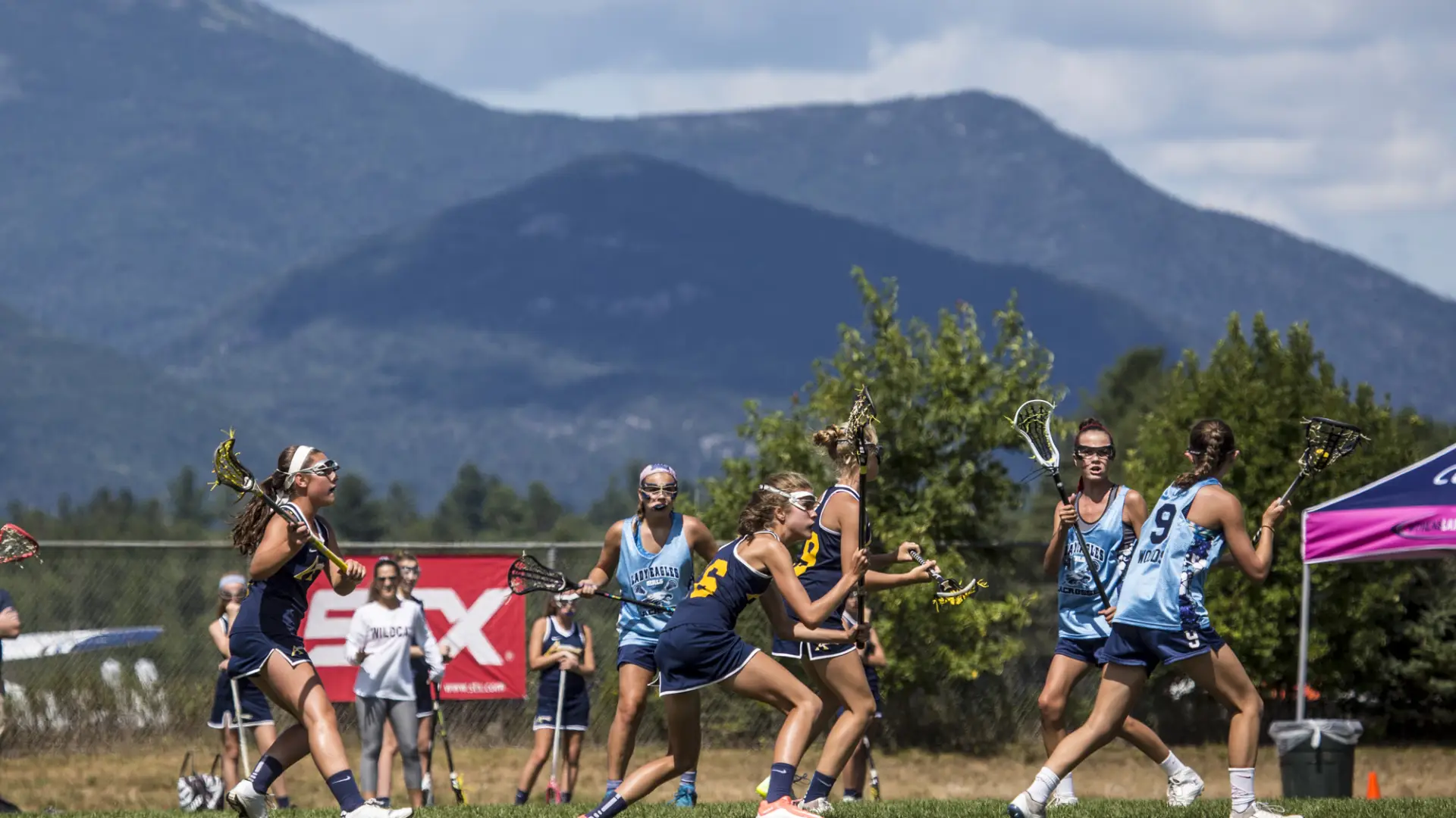 Two teams of young girls play lacrosse on green field with mountain backdrop.