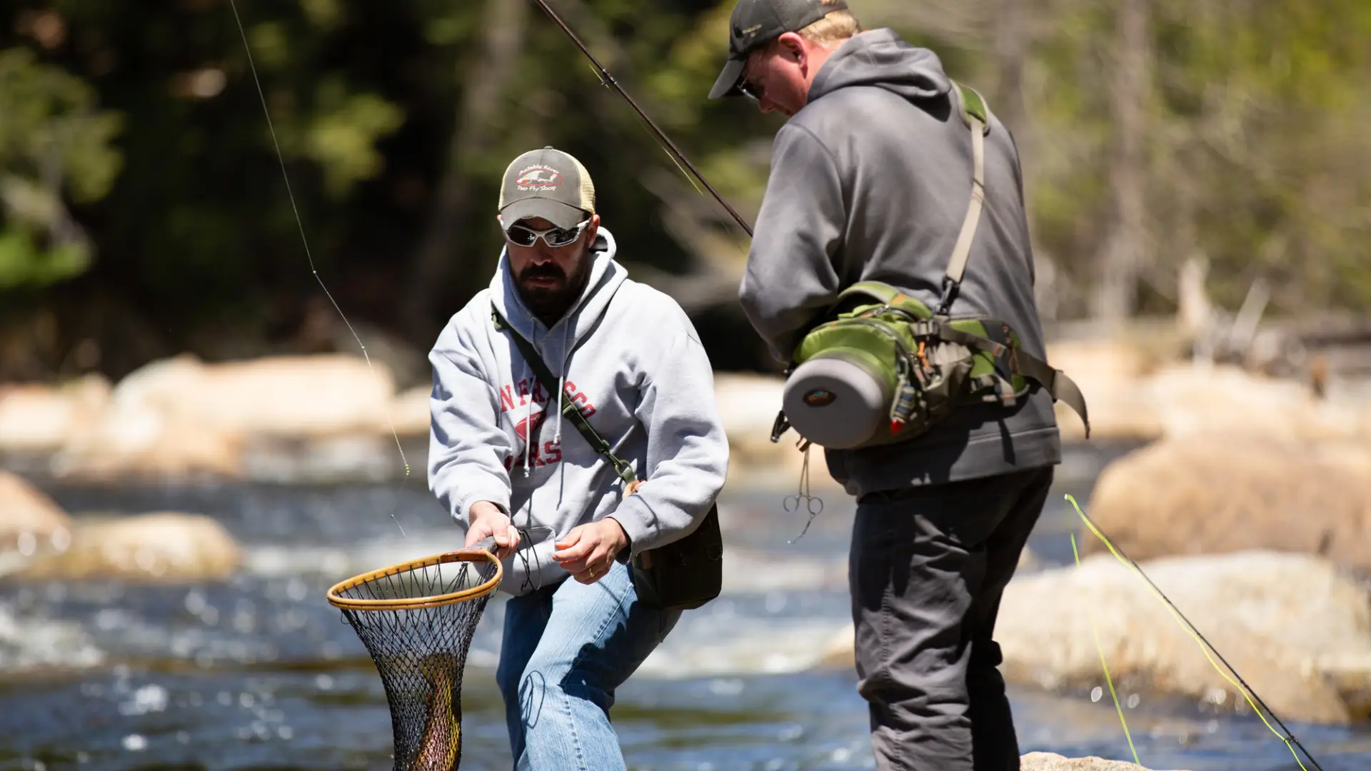 Two men fly fishing; one is reeling in a fish, while the other holds a net for the fish.