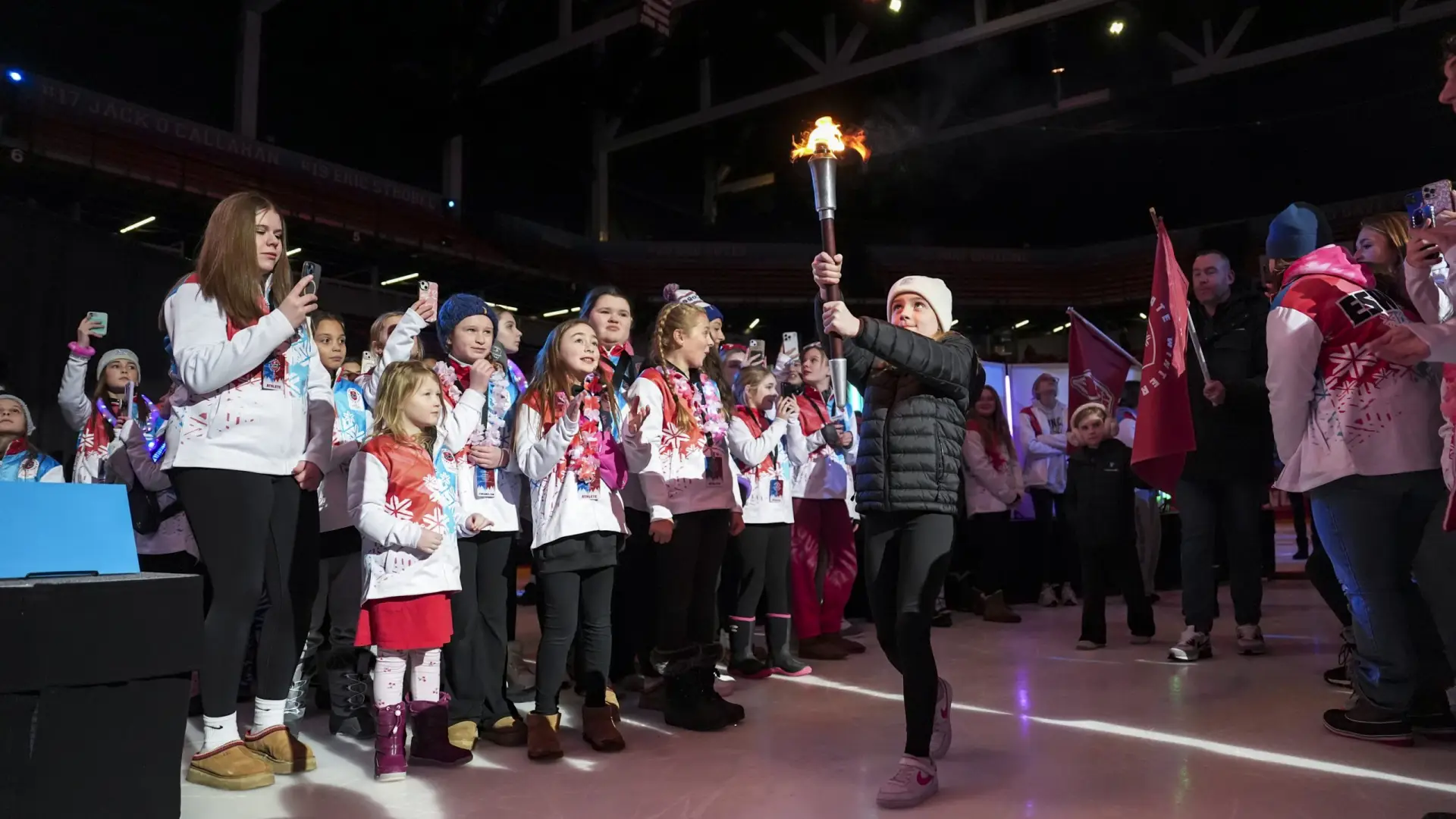 A young girl proudly carries a torch at the opening ceremony, surrounded by excited participants in ESWG jackets, capturing moments on their phones.