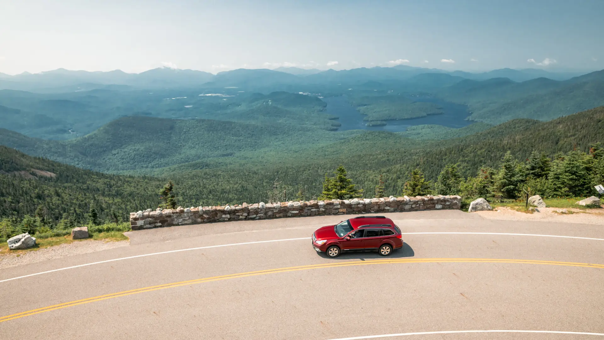 Red car drives on turn of high elevation highway road with mountain views and Lake Placid seen in the distance