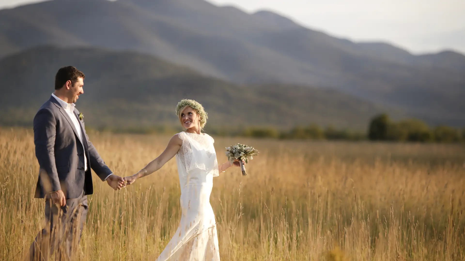 Bride and groom walk through a golden grass field with a mountain backdrop
