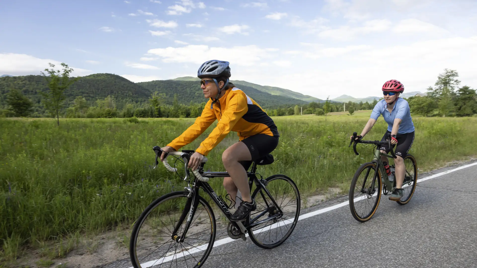 Cyclist wearing helmet and yellow and black rides single file ahead of second cyclist in light blue on paved road with green field and distant mountains in background